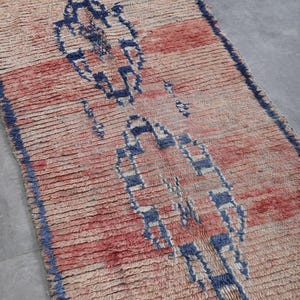May include: A vintage Moroccan rug with a geometric pattern in shades of blue, pink, and white. The rug has a distressed look and is made of wool.