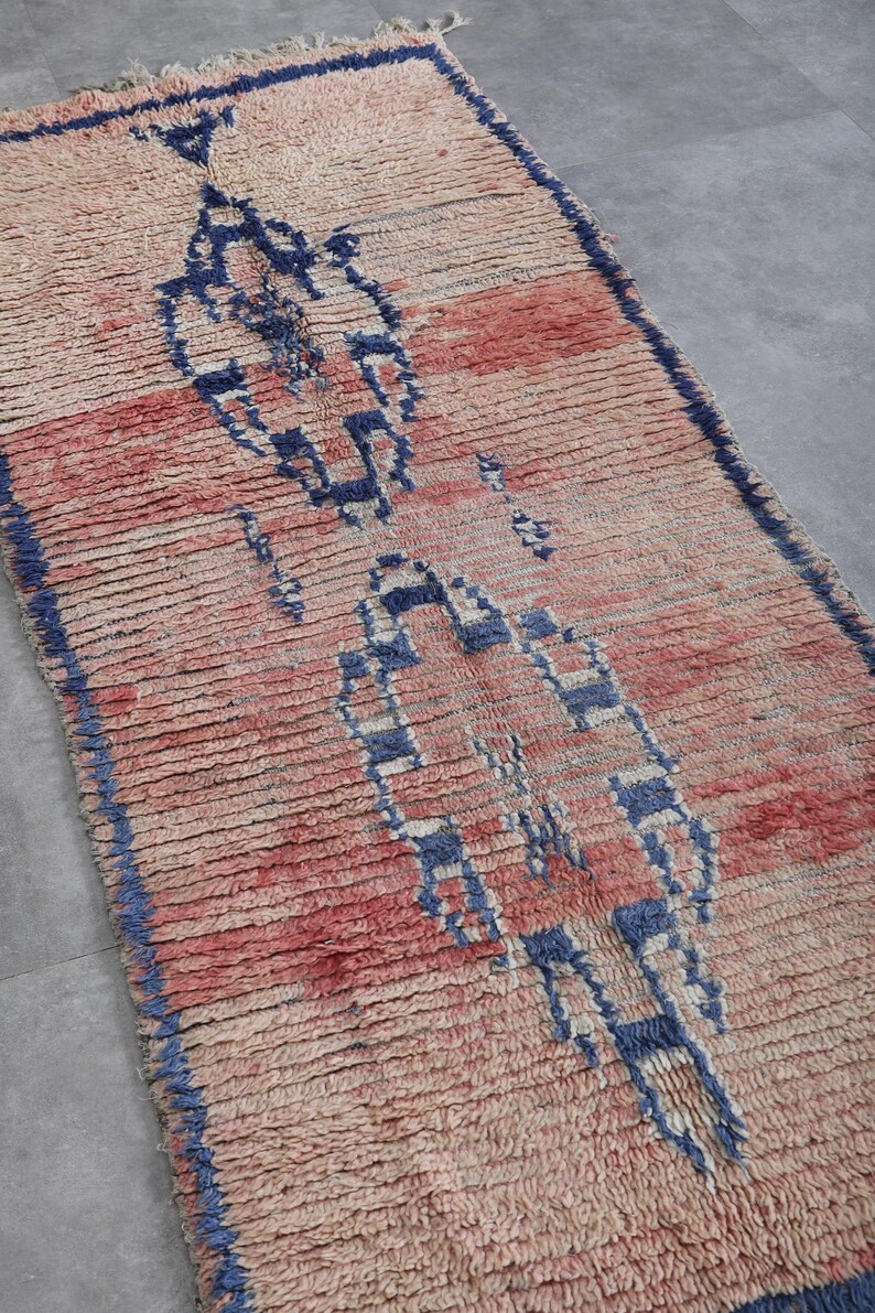 May include: A vintage Moroccan rug with a geometric pattern in shades of blue, pink, and white. The rug has a distressed look and is made of wool.