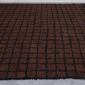 Peut inclure: Un tapis rectangulaire marron avec un motif quadrill&eacute; de lignes noires. Le tapis a une texture touffue et est pos&eacute; sur une surface gris clair. Le tapis est un article de d&eacute;coration int&eacute;rieure.