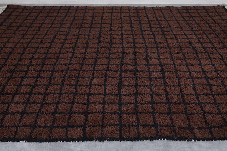 Peut inclure: Un tapis rectangulaire marron avec un motif quadrill&eacute; de lignes noires. Le tapis a une texture touffue et est pos&eacute; sur une surface gris clair. Le tapis est un article de d&eacute;coration int&eacute;rieure.