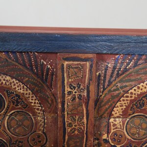 May include: Close-up of a section of a wooden chest with intricate hand-painted designs. The chest features a dark red base with gold, blue, and green accents. The top edge is a dark blue colour.