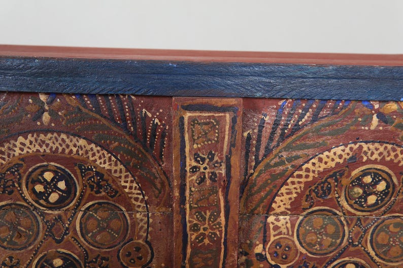 May include: Close-up of a section of a wooden chest with intricate hand-painted designs. The chest features a dark red base with gold, blue, and green accents. The top edge is a dark blue colour.