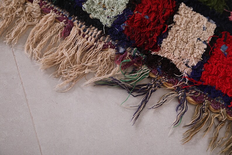 May include: Close-up of a colorful woven rug with fringe. The rug is made of different colored yarns, including red, blue, green, and white. The fringe is a light brown color.