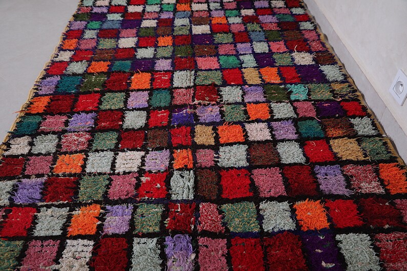 May include: A colorful Moroccan rug with a checkered pattern in shades of red, orange, green, purple, and black. The rug is made of wool and has a hand-woven texture.