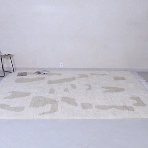 May include: A white shag rug with a beige abstract pattern. The rug has white fringe around the edges and is laid out on a light grey tiled floor.