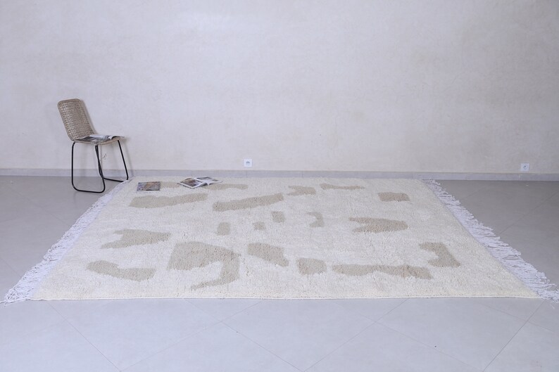 May include: A white shag rug with a beige abstract pattern. The rug has white fringe around the edges and is laid out on a light grey tiled floor.
