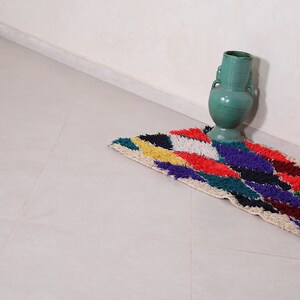 May include: A colorful, geometric patterned rug with a green ceramic vase in the corner of a room.