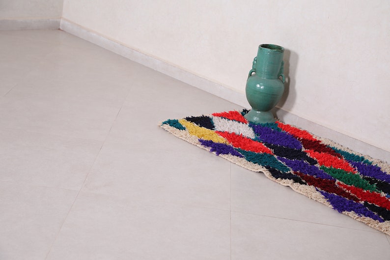 May include: A colorful, geometric patterned rug with a green ceramic vase in the corner of a room.