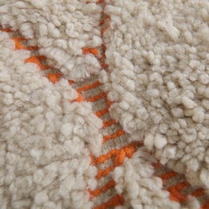 May include: Close-up of a cream-colored, textured rug with a thick pile. The rug features a pattern of orange lines running vertically and horizontally, creating a geometric design. The rug's texture appears soft and plush.