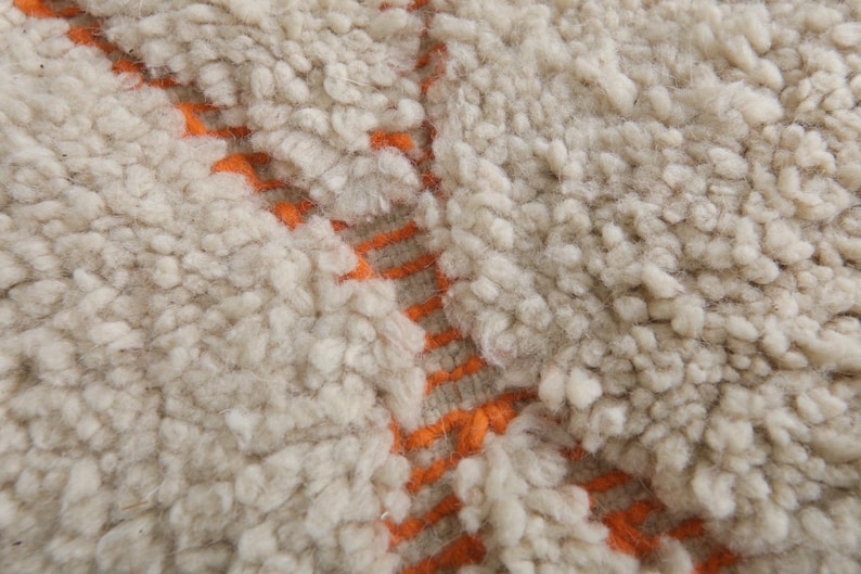 May include: Close-up of a cream-colored, textured rug with a thick pile. The rug features a pattern of orange lines running vertically and horizontally, creating a geometric design. The rug's texture appears soft and plush.