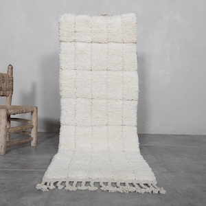 May include: A white, fluffy, rectangular rug with a fringe border. The rug is made of a soft, textured material and has a subtle pattern of squares.