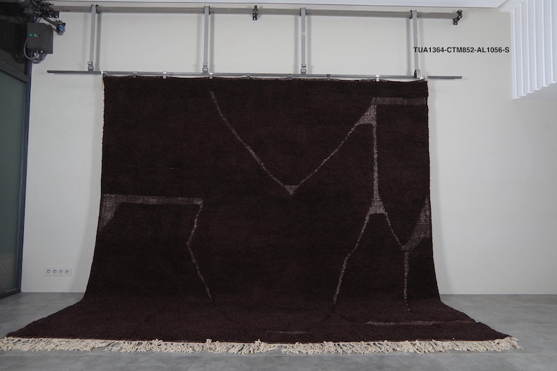 May include: A large, dark brown rug with a geometric design in lighter brown. The rug has a thick, textured pile and a fringe along the bottom edge. The rug is hanging from a metal bar against a white wall.