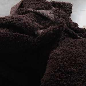 May include: A close-up of a dark brown, shaggy rug with a thick, textured pile. The rug is folded and layered, revealing a glimpse of a lighter, woven backing. The overall appearance suggests a soft, plush texture.