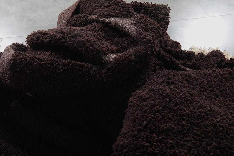 May include: A close-up of a dark brown, shaggy rug with a thick, textured pile. The rug is folded and layered, revealing a glimpse of a lighter, woven backing. The overall appearance suggests a soft, plush texture.