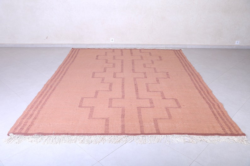 May include: A light pink rug with a geometric pattern in a darker shade of pink. The rug has a white fringe border.