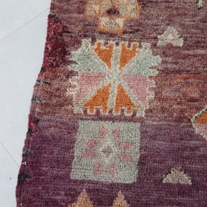 Może przedstawiać: Zbliżenie na kolorowy marokański dywan z misternymi geometrycznymi wzorami w odcieniach r&oacute;żowego, pomarańczowego i brązowego. Dywan ma ciemnobordowy kolor tła i fakturowany, ręcznie tkany wygląd.