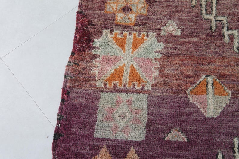 Może przedstawiać: Zbliżenie na kolorowy marokański dywan z misternymi geometrycznymi wzorami w odcieniach r&oacute;żowego, pomarańczowego i brązowego. Dywan ma ciemnobordowy kolor tła i fakturowany, ręcznie tkany wygląd.