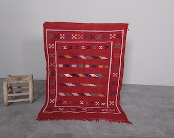Op de afbeelding: Een rood Marokkaans tapijt met een geometrisch patroon in tinten blauw, groen en wit. Het tapijt heeft een franje rand en is geweven met een traditioneel ontwerp.