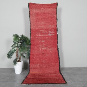 May include: A long, narrow red rug with a black border. The rug has a distressed, vintage look and is made of wool.