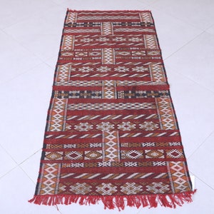 May include: A red, white, and black patterned rug with fringe. The rug has a geometric design with a Moroccan style.