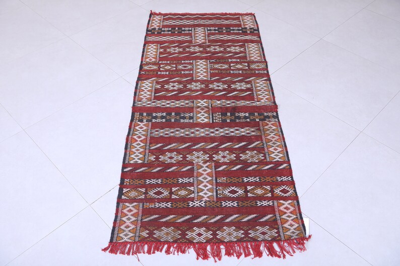 May include: A red, white, and black patterned rug with fringe. The rug has a geometric design with a Moroccan style.