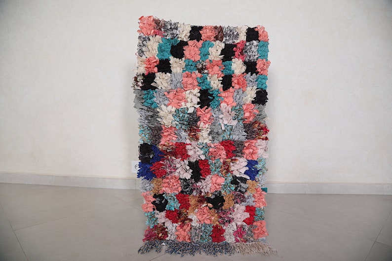 Puede incluir: Una alfombra colorida tejida a mano con un patr&oacute;n a cuadros en tonos de rosa, azul, gris y negro. La alfombra tiene un borde de flecos y est&aacute; hecha de materiales reciclados.
