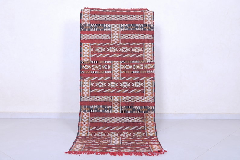 May include: A red, white, and black patterned rug with geometric designs. The rug has a fringe on the bottom.