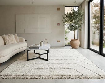 May include: A modern living room featuring a cream-colored sofa with fluffy pillows, a marble coffee table, and a large, textured white rug with geometric patterns. An olive tree in a terracotta pot sits near a large window.