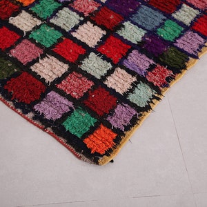 May include: A colorful rug with a patchwork design featuring squares of various colors, including red, green, purple, and orange. The rug is made of a thick, textured material and has a fringe border.