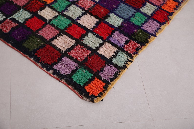May include: A colorful rug with a patchwork design featuring squares of various colors, including red, green, purple, and orange. The rug is made of a thick, textured material and has a fringe border.