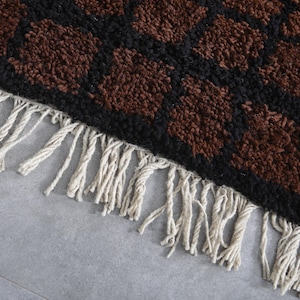 Peut inclure: Gros plan sur un tapis avec un motif r&eacute;p&eacute;titif de formes marron sur un fond noir. Le tapis a une frange de glands cr&egrave;me le long d'un bord. Le tapis est probablement fait d'une mati&egrave;re douce et textur&eacute;e.