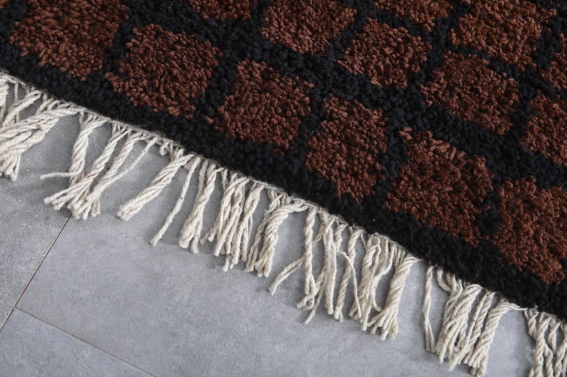 Peut inclure: Gros plan sur un tapis avec un motif r&eacute;p&eacute;titif de formes marron sur un fond noir. Le tapis a une frange de glands cr&egrave;me le long d'un bord. Le tapis est probablement fait d'une mati&egrave;re douce et textur&eacute;e.