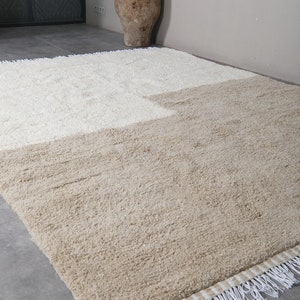 Puede incluir: Una alfombra de pelo beige y blanco con flecos. La alfombra est&aacute; colocada sobre un suelo de baldosas grises.