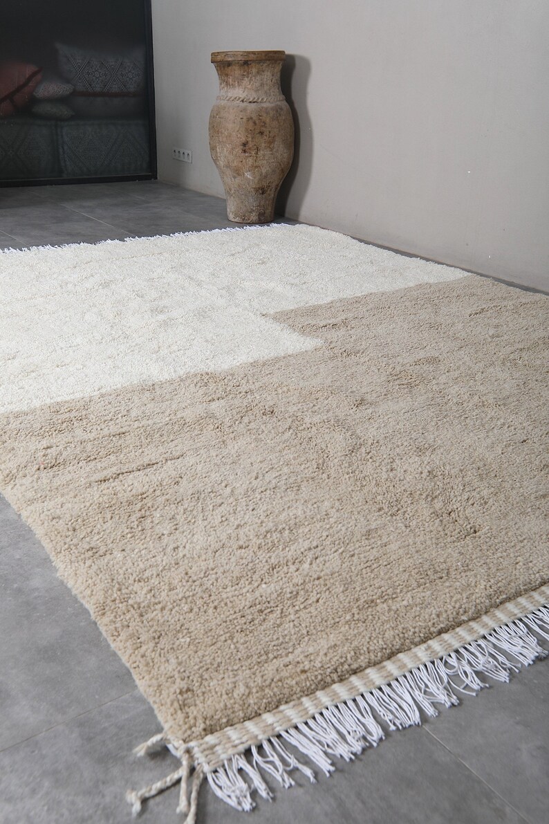 Puede incluir: Una alfombra de pelo beige y blanco con flecos. La alfombra est&aacute; colocada sobre un suelo de baldosas grises.