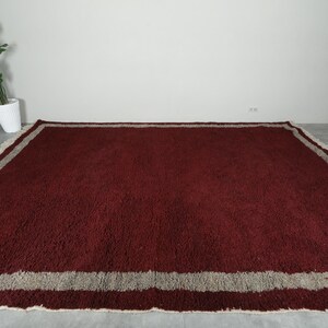 May include: A large, rectangular rug with a deep burgundy field and a wide, light gray border. The rug has a shaggy texture and a fringed edge, suitable for a living room or bedroom.