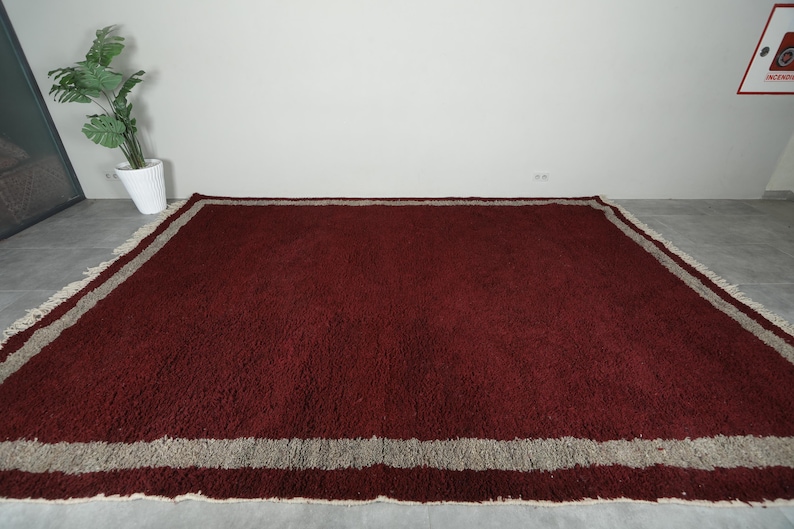 May include: A large, rectangular rug with a deep burgundy field and a wide, light gray border. The rug has a shaggy texture and a fringed edge, suitable for a living room or bedroom.