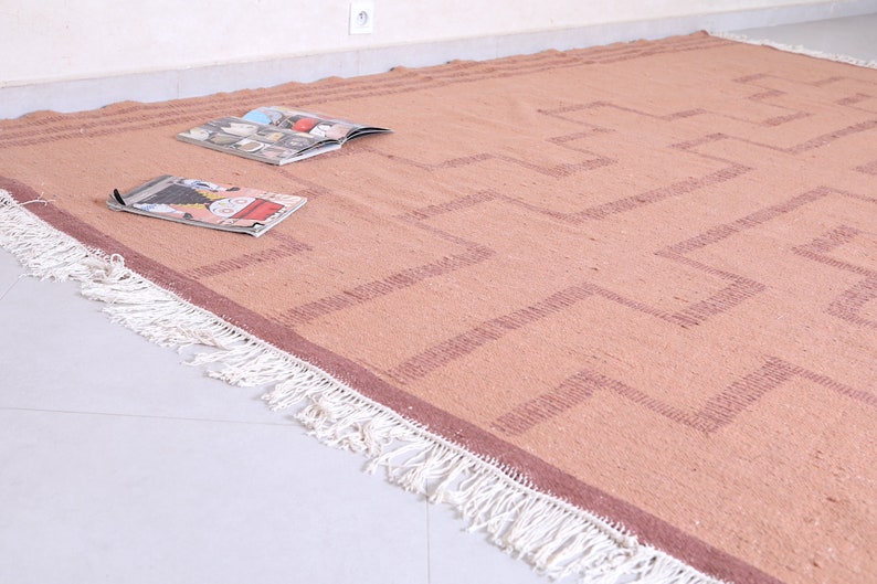 May include: A light brown rug with a geometric pattern in a darker brown colour. The rug has white fringe around the edges.