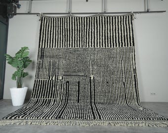 May include: A large, black and white striped rug with a fringe along the bottom edge. The rug has a geometric pattern with vertical stripes and rectangular shapes. The rug is displayed against a neutral wall.