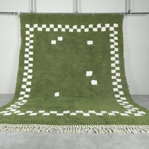 May include: A green rug with a white checkered border and scattered white squares. The rug has a shaggy texture and a fringe along the bottom edge. The rug is hanging from a metal bar.