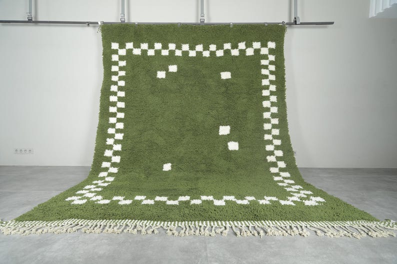 May include: A green rug with a white checkered border and scattered white squares. The rug has a shaggy texture and a fringe along the bottom edge. The rug is hanging from a metal bar.