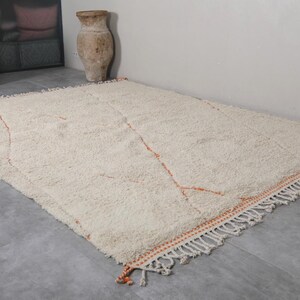 May include: A large, rectangular, off-white rug with a shaggy texture. The rug features a simple design with thin, orange lines and a fringed border. The rug is placed on a gray floor, and the overall aesthetic is neutral and inviting.
