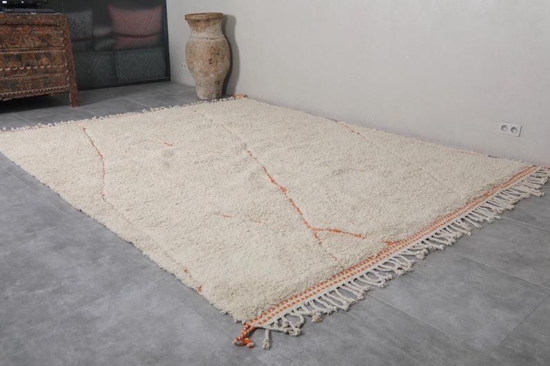 May include: A large, rectangular, off-white rug with a shaggy texture. The rug features a simple design with thin, orange lines and a fringed border. The rug is placed on a gray floor, and the overall aesthetic is neutral and inviting.