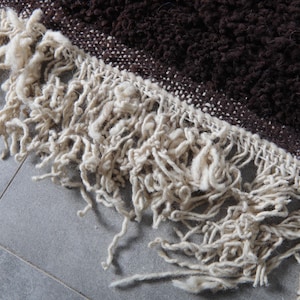 May include: Close-up of a brown and cream-colored rug. The rug has a textured, shaggy appearance with a fringe of cream-colored yarn. The brown portion of the rug is a darker shade, and the fringe is a lighter, neutral tone.
