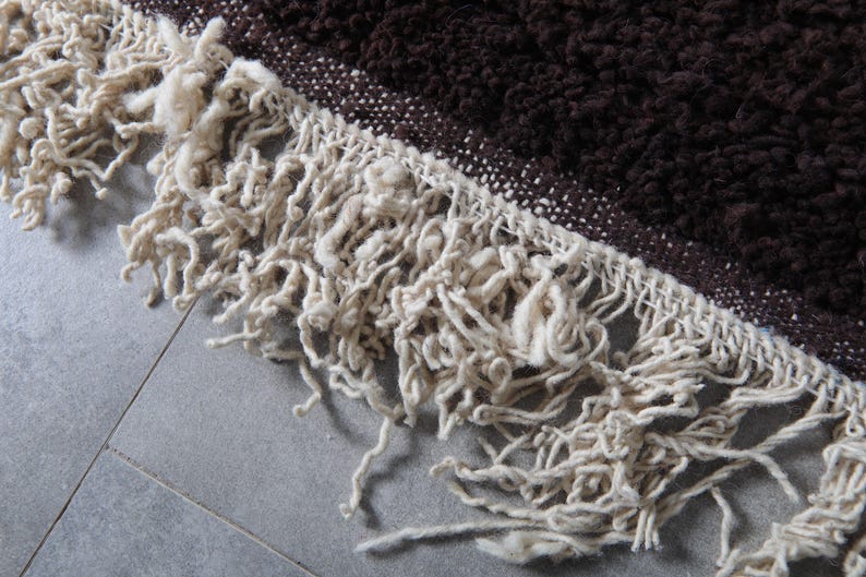 May include: Close-up of a brown and cream-colored rug. The rug has a textured, shaggy appearance with a fringe of cream-colored yarn. The brown portion of the rug is a darker shade, and the fringe is a lighter, neutral tone.