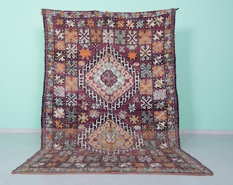 Puede incluir: Una alfombra marroquí vintage con un patrón geométrico en tonos de morado, naranja y crema. La alfombra tiene una forma de diamante central con detalles intrincados y un borde de formas geométricas más pequeñas.