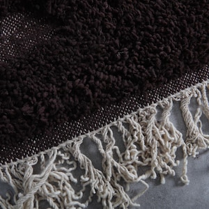 May include: Close-up of a dark brown, textured rug with a cream-colored fringe. The rug has a woven border and a plush, shaggy pile. The fringe is made of thick, twisted yarn.