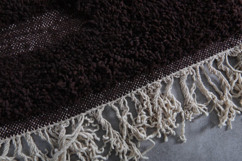 May include: Close-up of a dark brown, textured rug with a cream-colored fringe. The rug has a woven border and a plush, shaggy pile. The fringe is made of thick, twisted yarn.