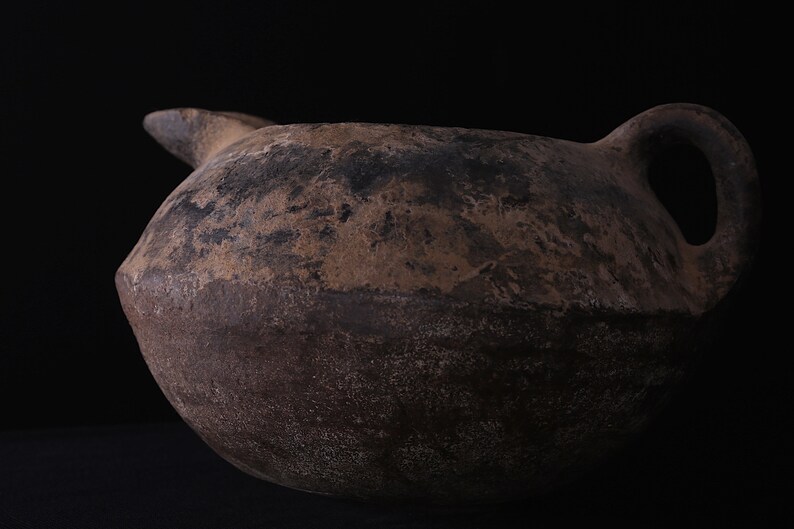 May include: A large, brown, earthenware jug with a wide, rounded body and a single handle. The jug has a rough, textured surface and appears to be very old.