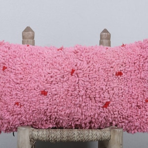 May include: A pink cushion with red accents. The cushion is made of a soft, fluffy material and has a slightly textured surface.