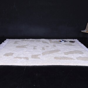 May include: White shag rug with a geometric pattern in a lighter shade of beige. The rug has a fringe border and is photographed on a black surface.